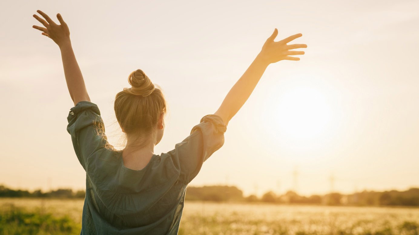 Junge Frau streckt auf einem Feld im Gegenlicht beide Arme in den Himmel