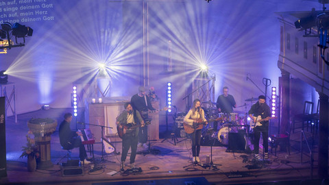 Band musiziert auf einer Bühne in einer Kirche in blau-violettem Licht, Liedtext wird an die Wand projiziert