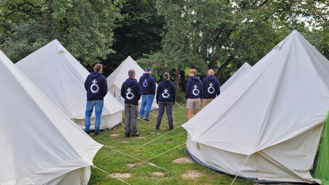 Sechs Teamer in dunkelblauen Hoodies stehen zwischen weißen Glockenzelten auf einer Wiese